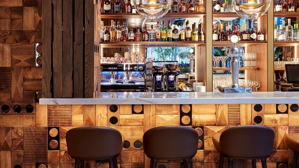 Uma área de bar com painéis de madeira, banquetas e diversos tipos de garrafas de bebidas.