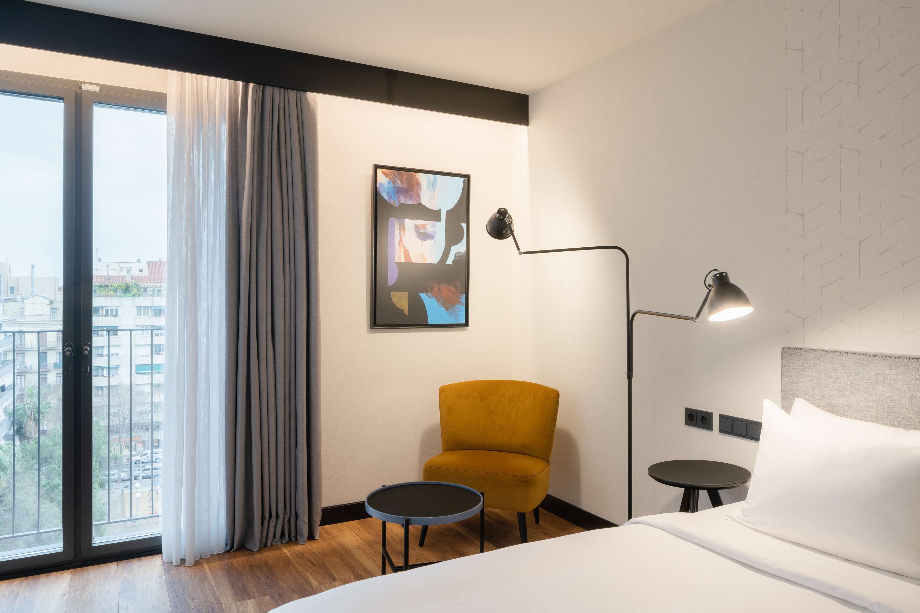Habitación de hotel con cama, una pequeña mesa redonda, un sillón amarillo, una lámpara de pie y un amplio ventanal con cortinas.
