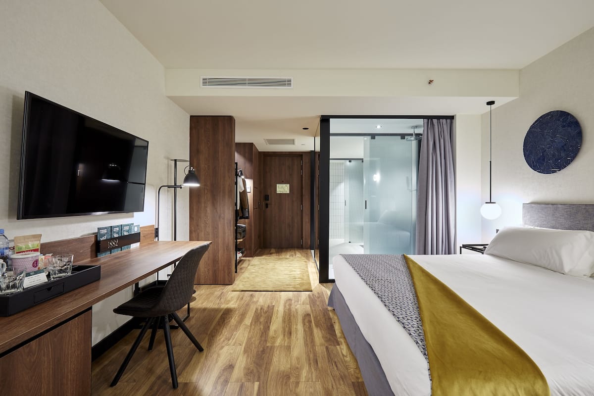 A modern hotel room with a flat-screen TV, a wooden desk with a chair, a bed with a gold throw, and a large round wall decoration.