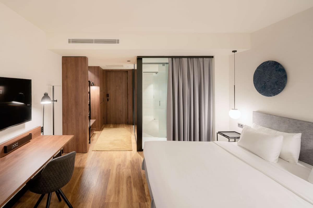 A modern hotel room with a large bed, a desk with a chair, a television, and a bathroom visible through a glass door.
