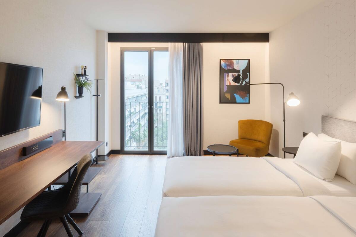 A hotel room with a bed, a desk, a chair, a TV, a lamp, and a window with curtains.