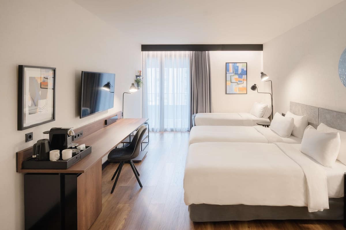 A hotel room with a wooden desk, a flat-screen TV, two beds, a small table, and a large window with curtains.