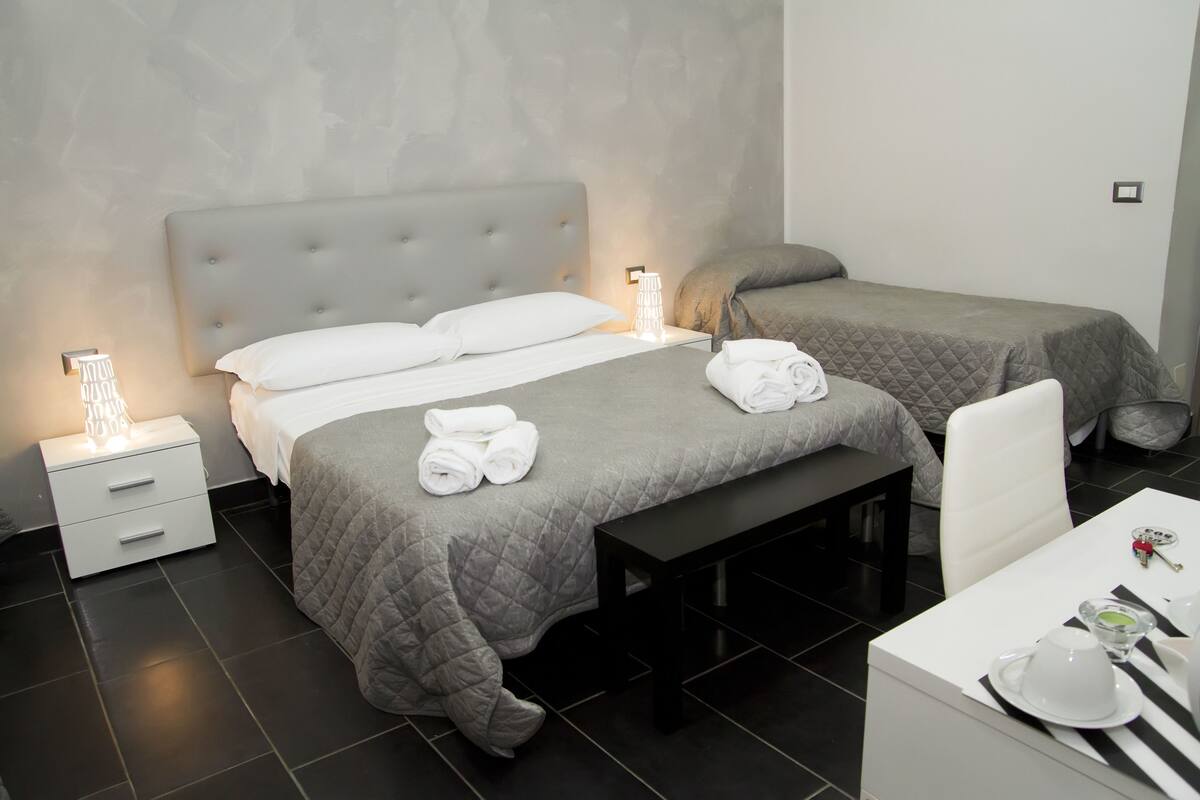 A hotel room with a large bed, a smaller bed, a bench, a desk with a chair, and a bedside table with a lamp.
