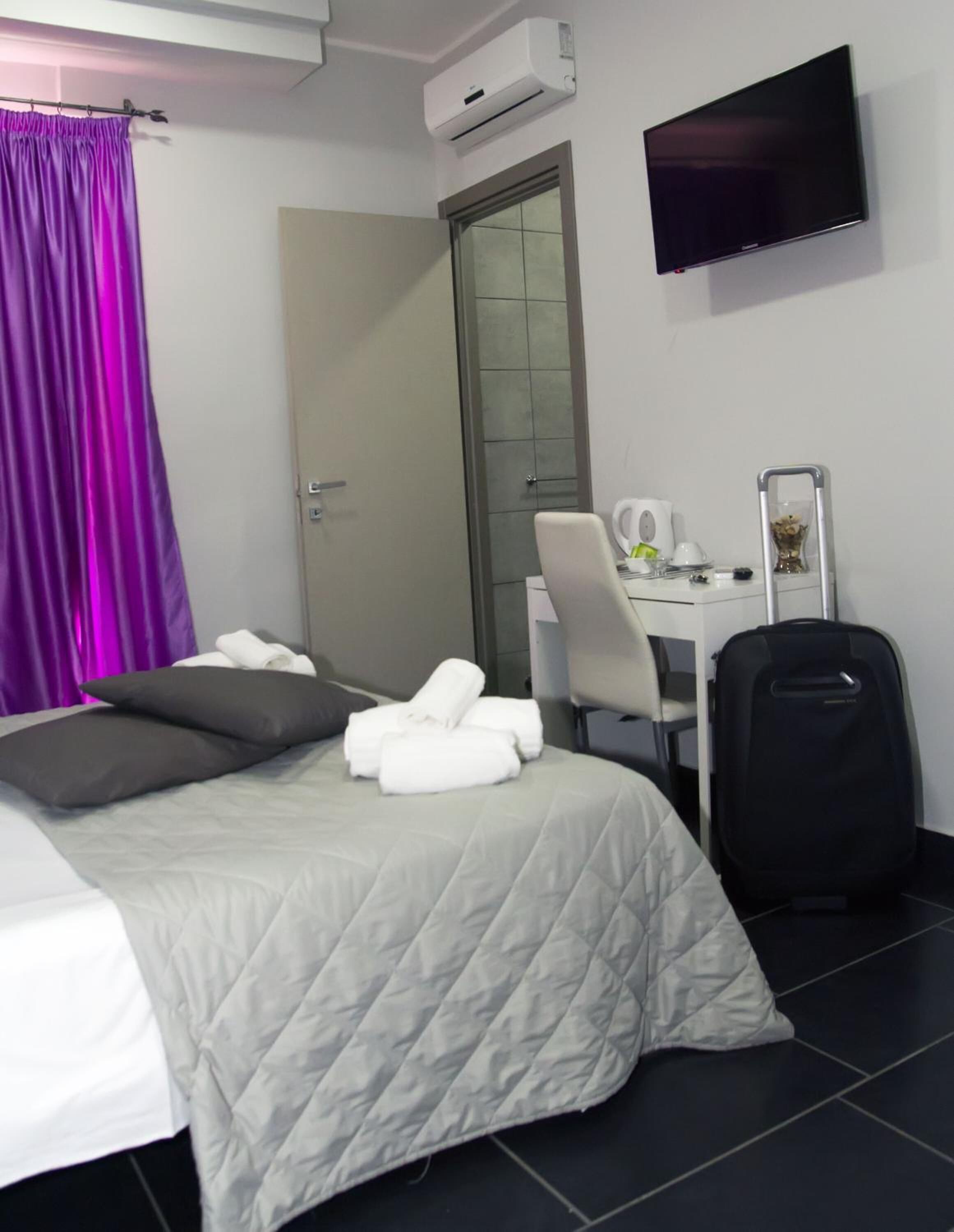 A hotel room with a bed, a desk with a chair, a suitcase, and a TV on the wall.