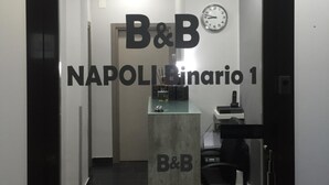 Zona de recepción con un mostrador, sillas, un reloj y un cartel que dice "B&B NAPOLI Binario 1".