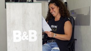 Una persona sentada en un escritorio con un letrero de B&B, sosteniendo documentos, en una habitación con una televisión de pantalla plana y una planta al fondo.