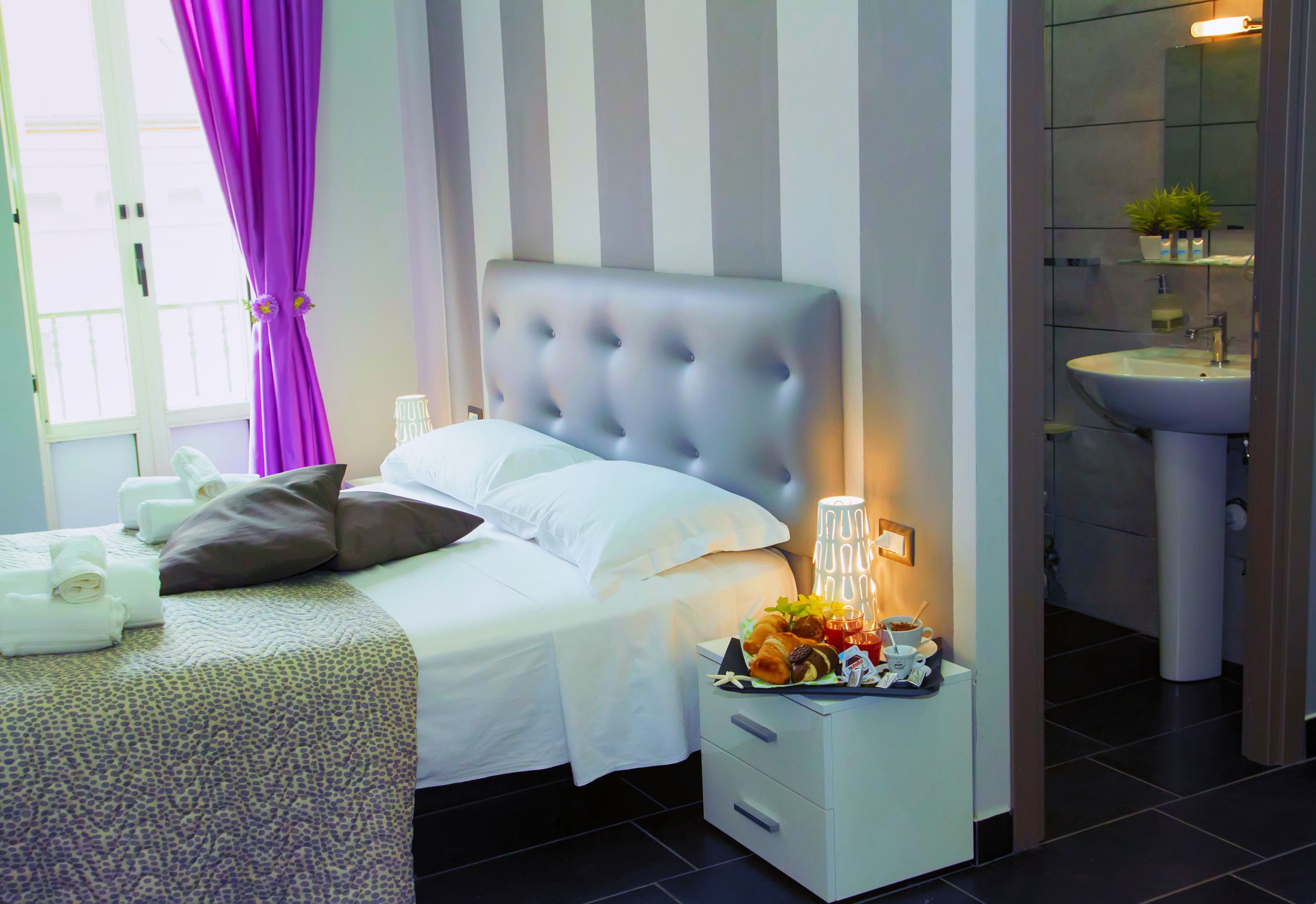 A neatly made bed with white linens and a tufted headboard, a bedside table with a lamp and a fruit basket, and a bathroom with a sink and mirror.