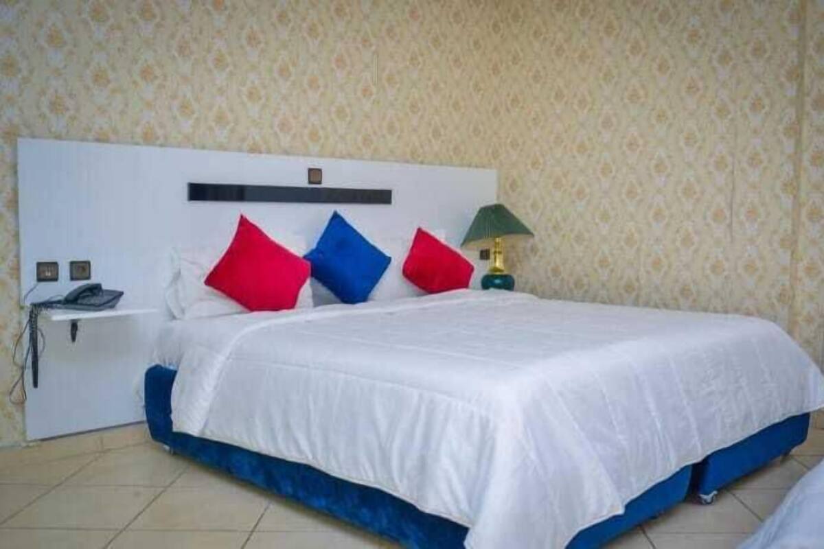A neatly made bed with a white and blue bedspread, a headboard with a shelf, and a nightstand with a lamp and phone.