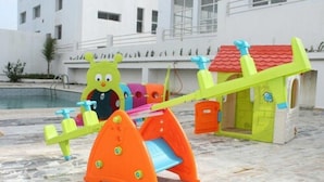 Un parque infantil con un balancín y un tobogán, frente a un edificio de techo rojo.