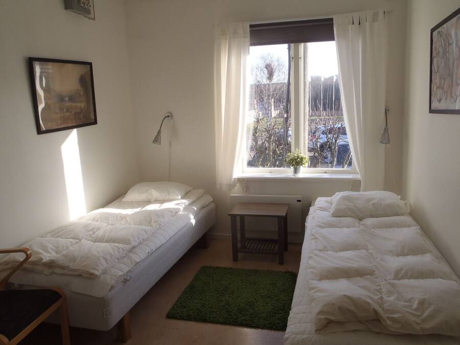 Una stanza con due letti singoli, una piccola scrivania, una sedia, una finestra vista alberi e un quadro appeso al muro.