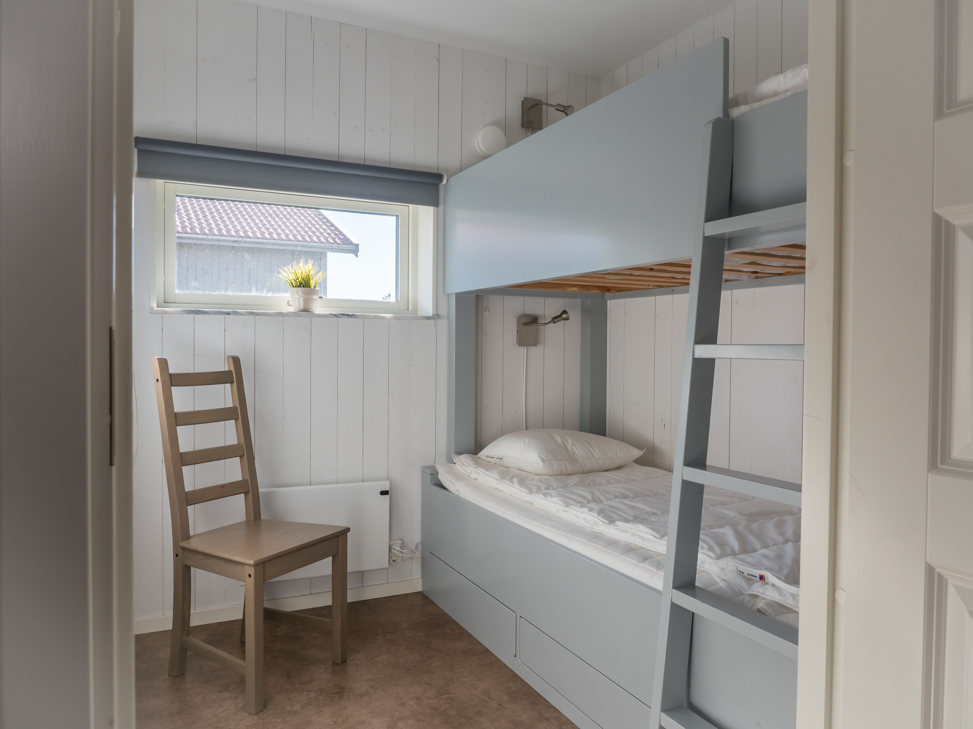 Ett sovrum med en våningssäng, en stol, ett fönster med utsikt över ett hus och ett element.