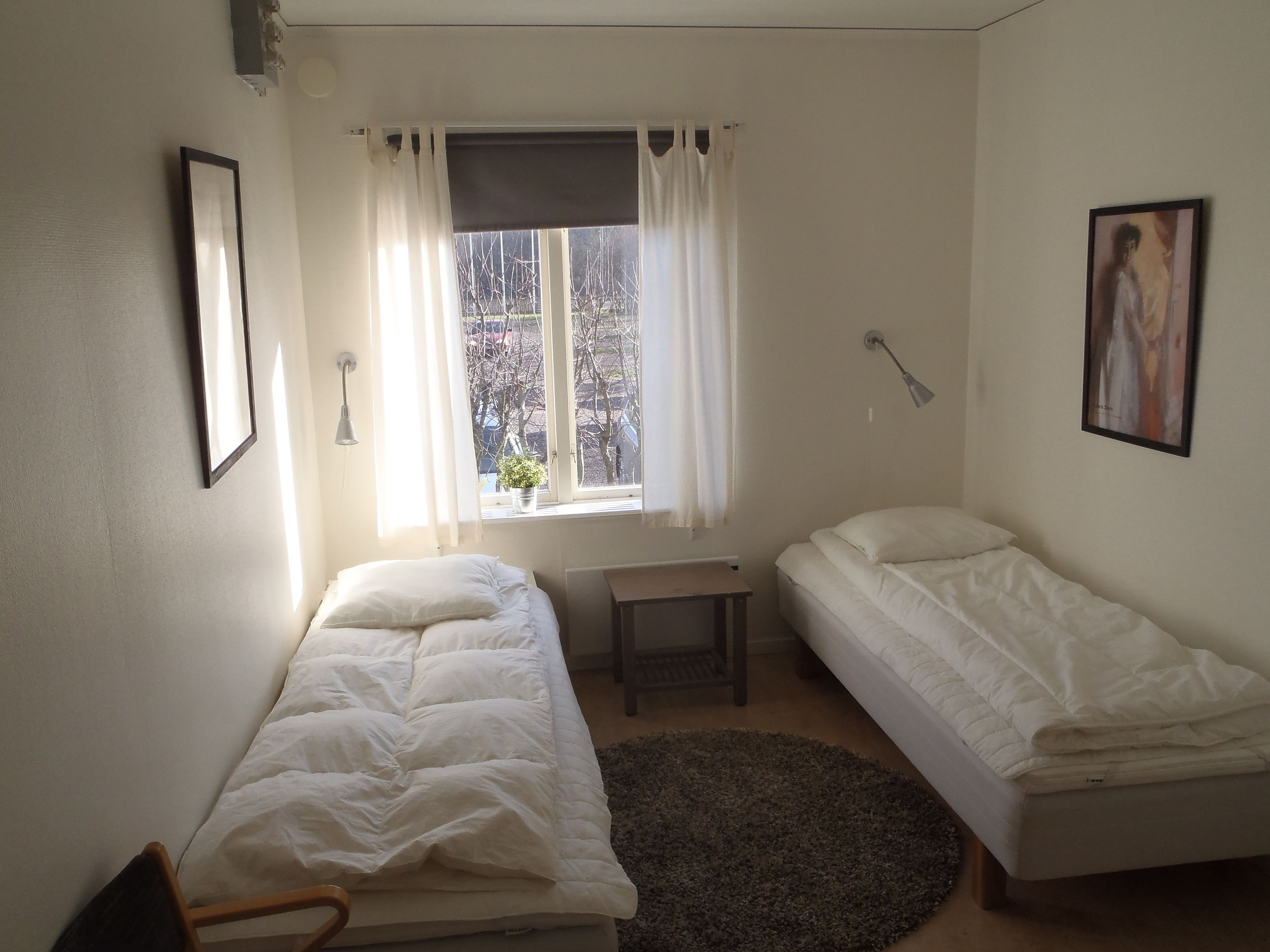 A small, well-lit room with two single beds, a small table, a chair, a window with curtains, and a framed picture on the wall.