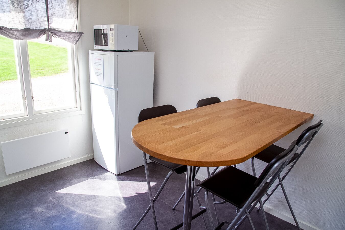Een klein eetgedeelte met een houten tafel en vier stoelen, een witte koelkast, en een raam met uitzicht op een gazon.