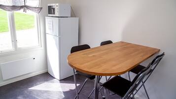 Een klein eetgedeelte met een houten tafel en vier stoelen, een witte koelkast, en een raam met uitzicht op een gazon.