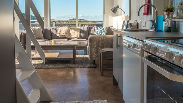 A modern kitchen with a large window, a dining area with a sofa, and a staircase leading to an upper level. - Hav & Logi Skärhamn (Skaerhamn)