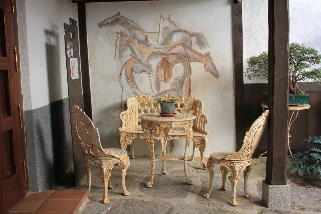 Un espace extérieur aménagé avec des chaises et une table décorées, un mur orné d’un tableau représentant un cheval et une plante en pot.