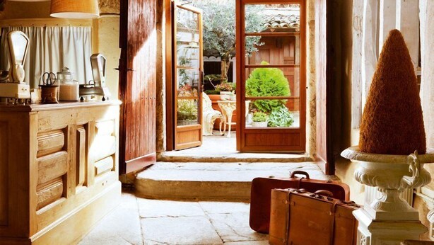 Un interior rústico con suelo de piedra, muebles de madera y vistas a un jardín exterior a través de una puerta abierta.