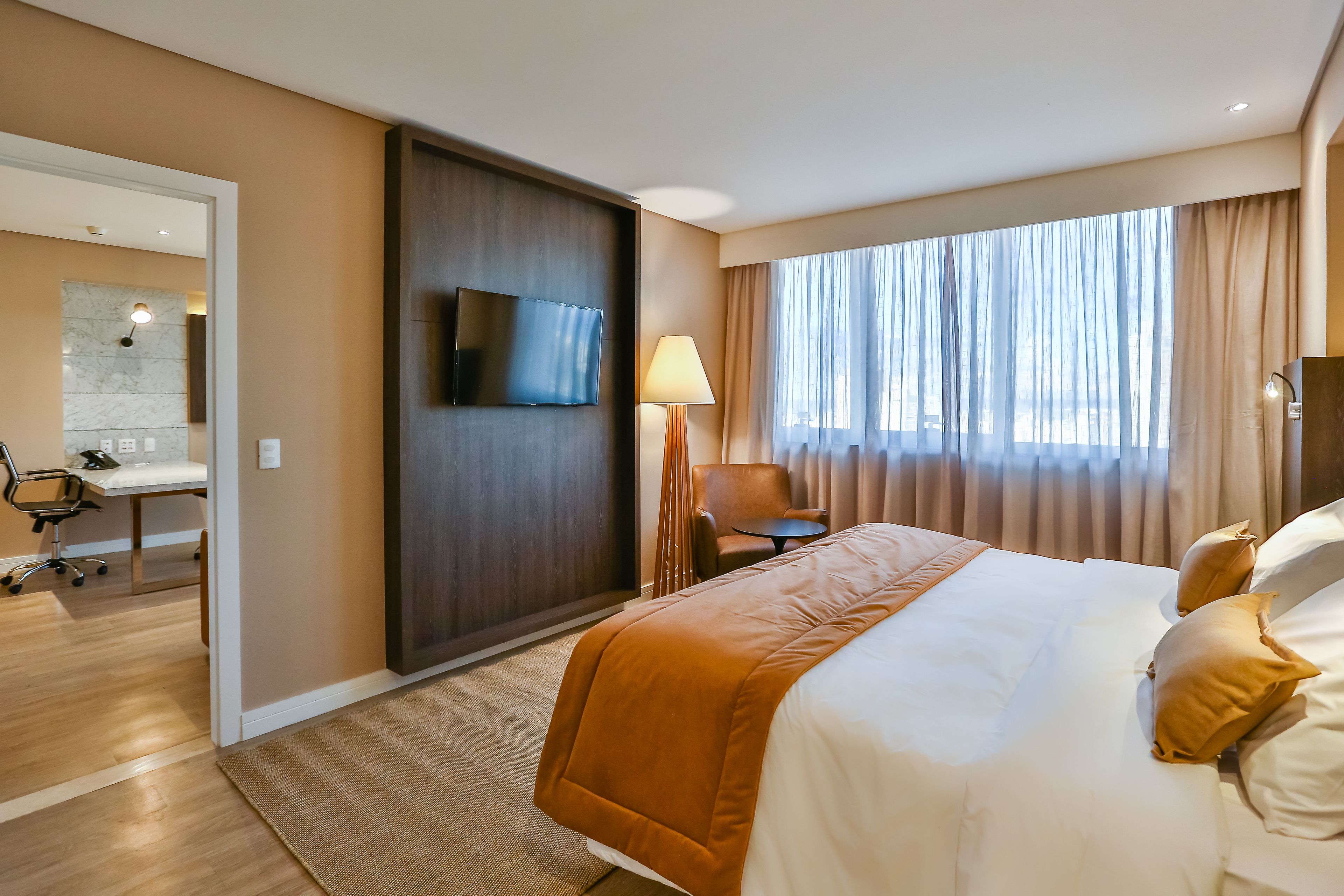 A hotel room with a large bed, a desk, a chair, a TV, and a window with curtains.
