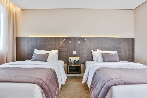 Habitación de hotel con dos camas, cabecero de madera, mesitas de noche con lámparas y una pequeña mesita.