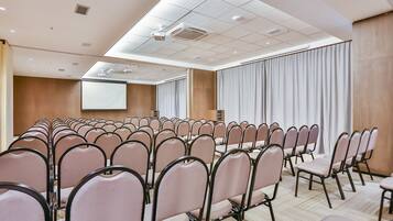 Uma sala de conferências com fileiras de cadeiras, uma tela de projetor e um grande janelão com cortinas.