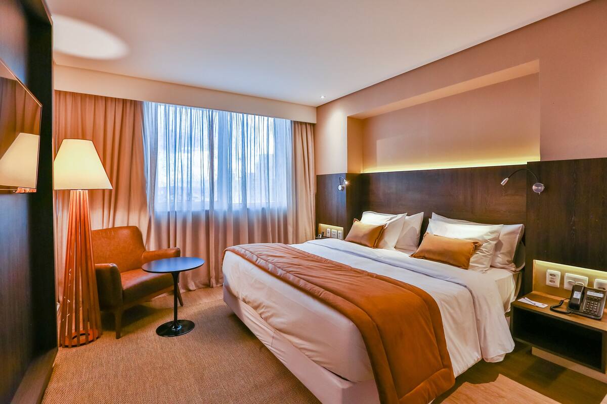 Una camera d'hotel con un letto grande, una sedia, un tavolino, un comodino con il telefono e una finestra con tende.