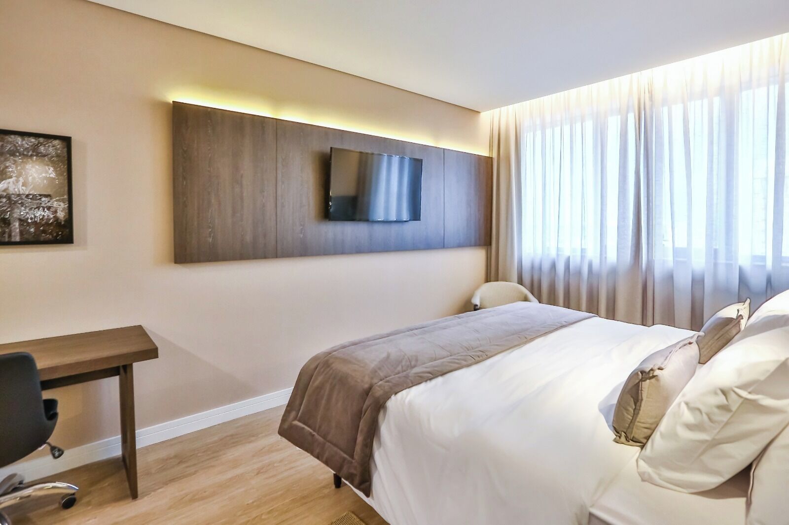 A hotel room with a bed, a desk, a chair, a flat-screen TV mounted on the wall, and a window with curtains.