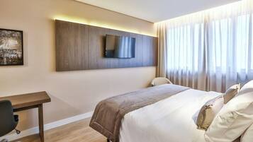 A hotel room with a bed, a desk, a chair, a flat-screen TV mounted on the wall, and a window with curtains.