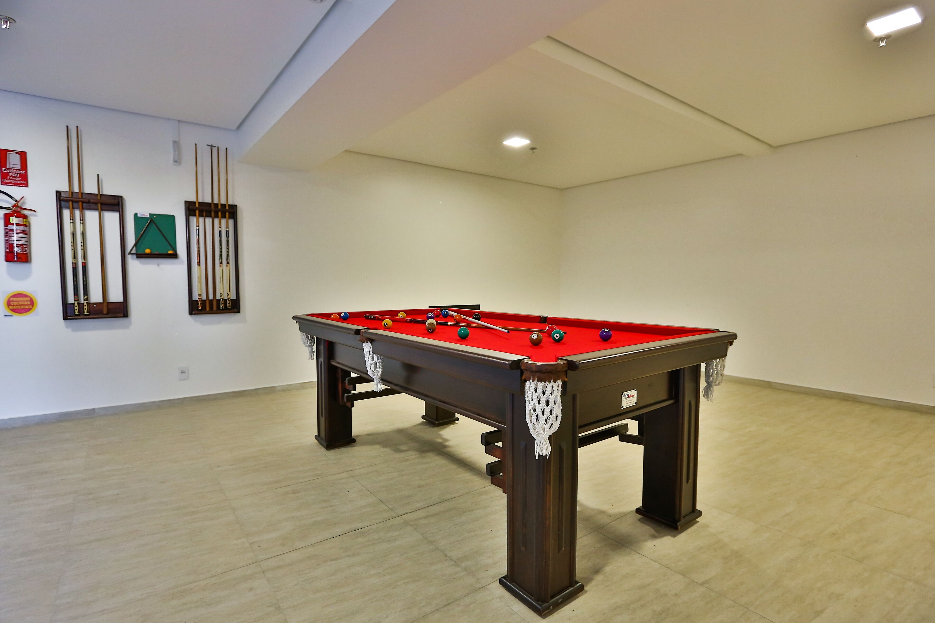 A pool table with a red felt surface, surrounded by walls with mounted cue sticks and a fire extinguisher.