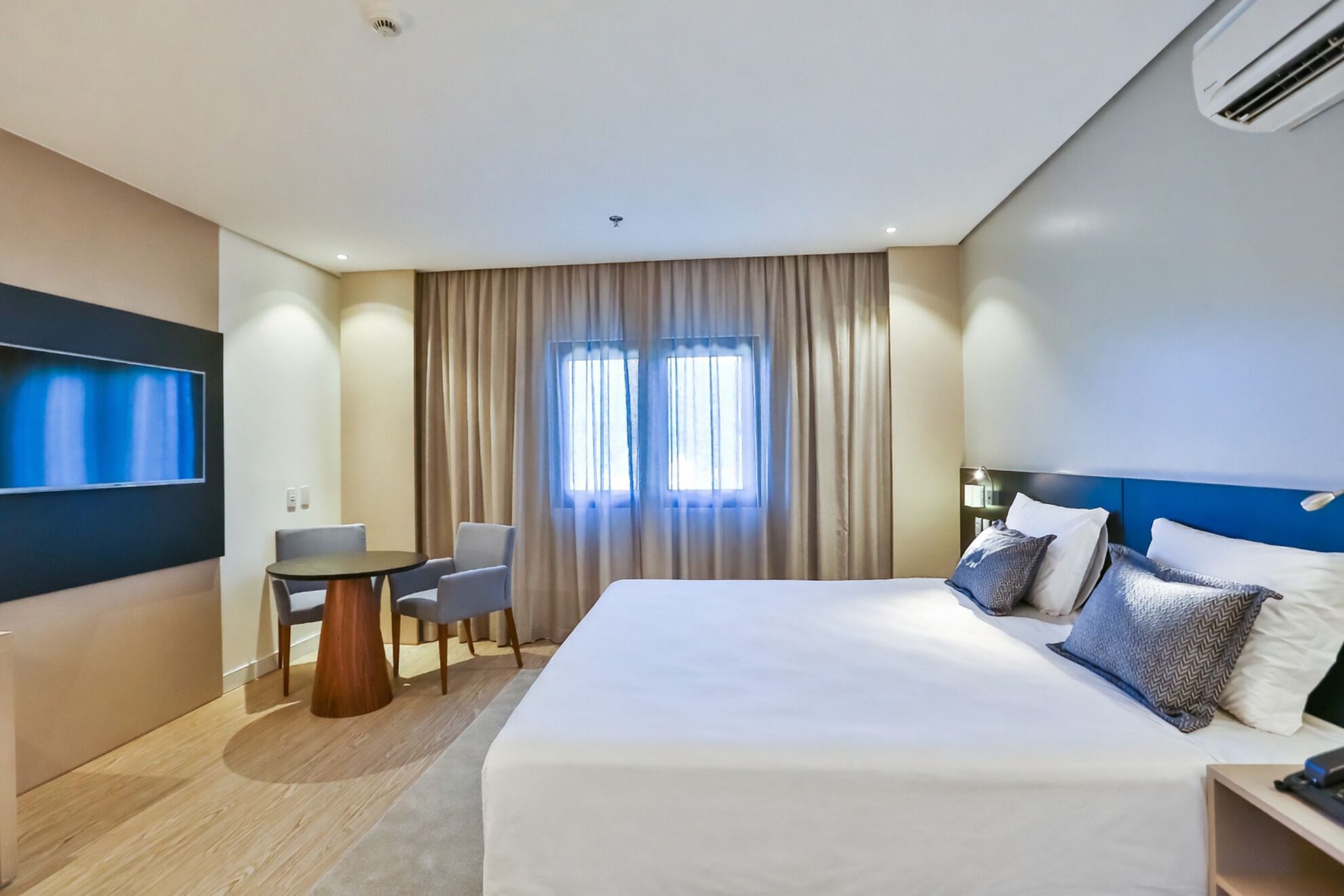 Quarto de hotel com uma cama grande, uma mesa redonda pequena com duas cadeiras, uma televisão presa na parede e uma janela com cortinas.