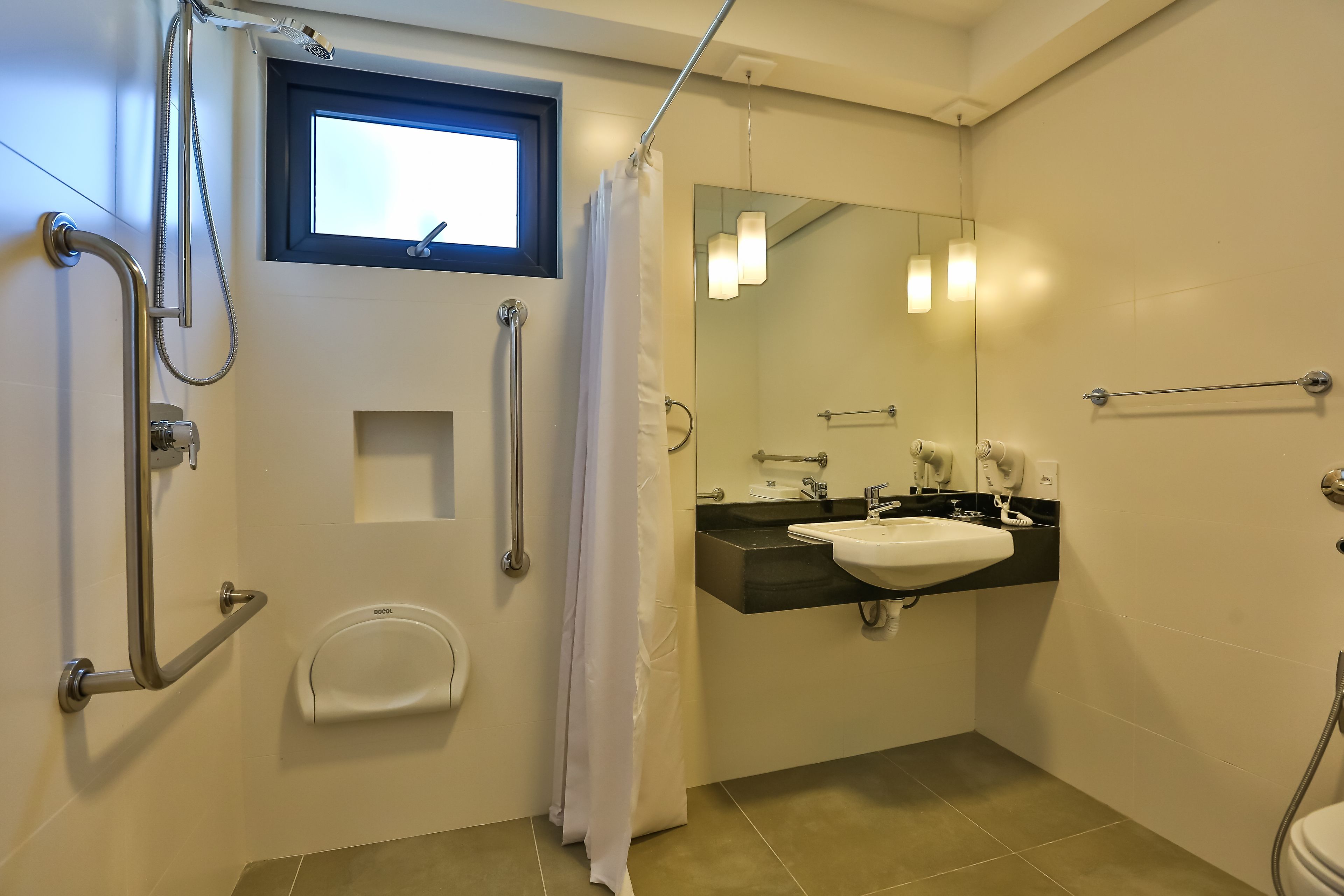 A bathroom with a shower, toilet, and sink. It includes grab bars, a window, and a mirror.