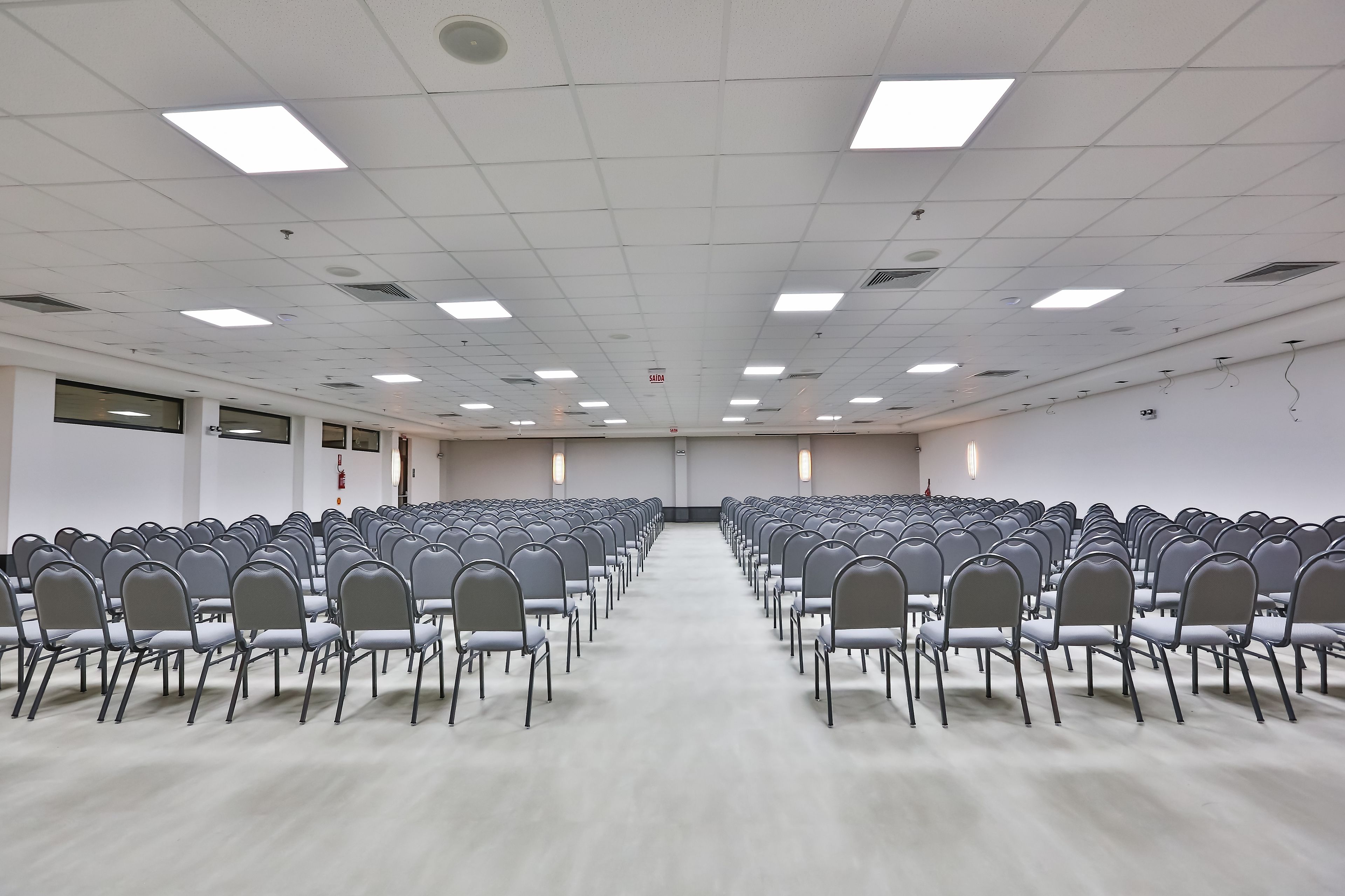 Un salón de conferencias con filas de sillas, un techo blanco con luces cuadradas y ventanas amplias a lo largo de una pared.
