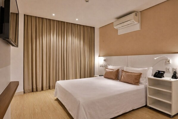 A hotel room with a large bed, a bedside table with a phone and a lamp, a wall-mounted air conditioner, and a curtain.