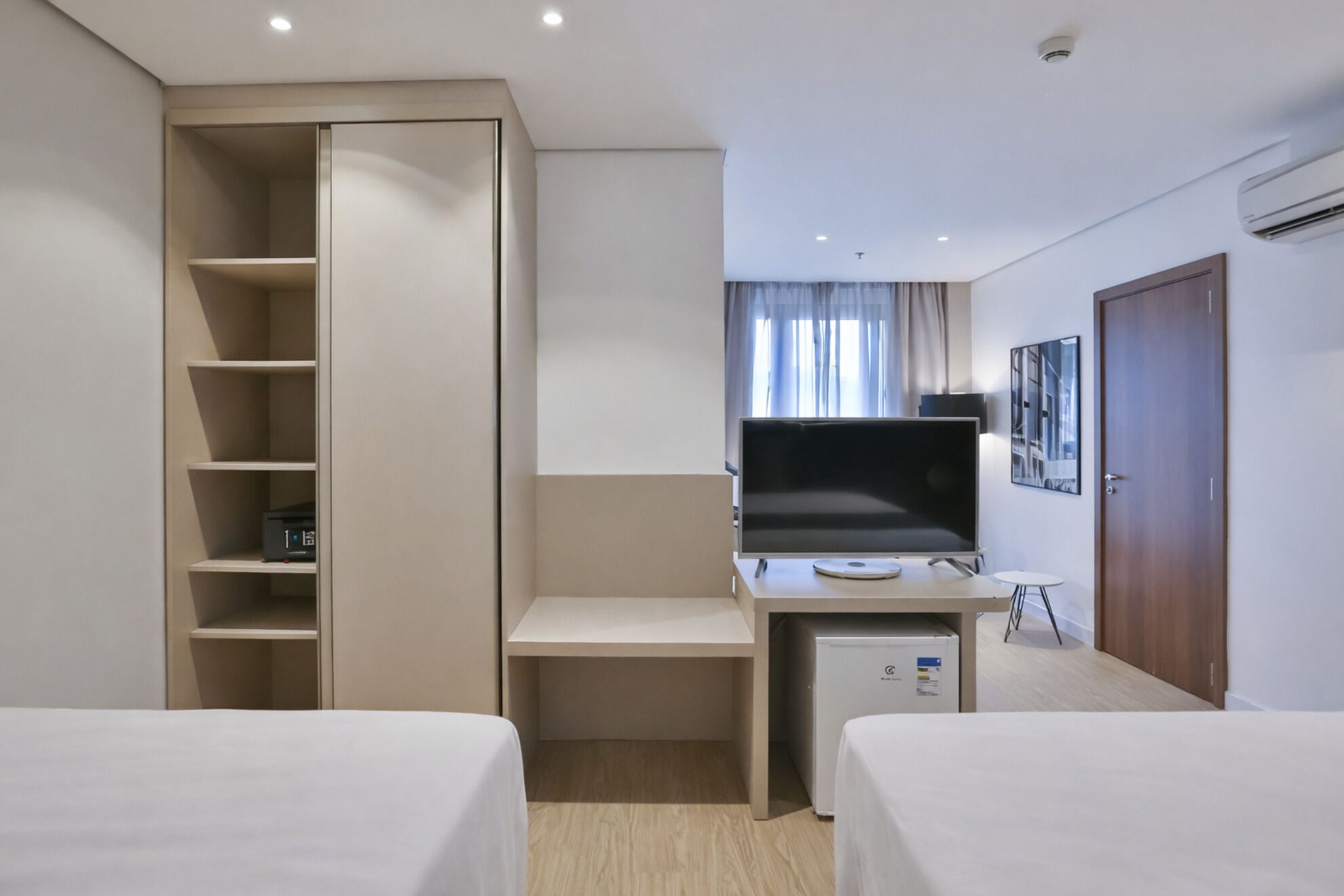 A hotel room with two beds, a desk with a computer, a TV, a wardrobe, and an air conditioning unit.