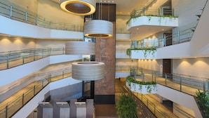 Un atrio de varios niveles con arquitectura moderna, que presenta luminarias cilíndricas, barandillas de vidrio y plantas verdes.