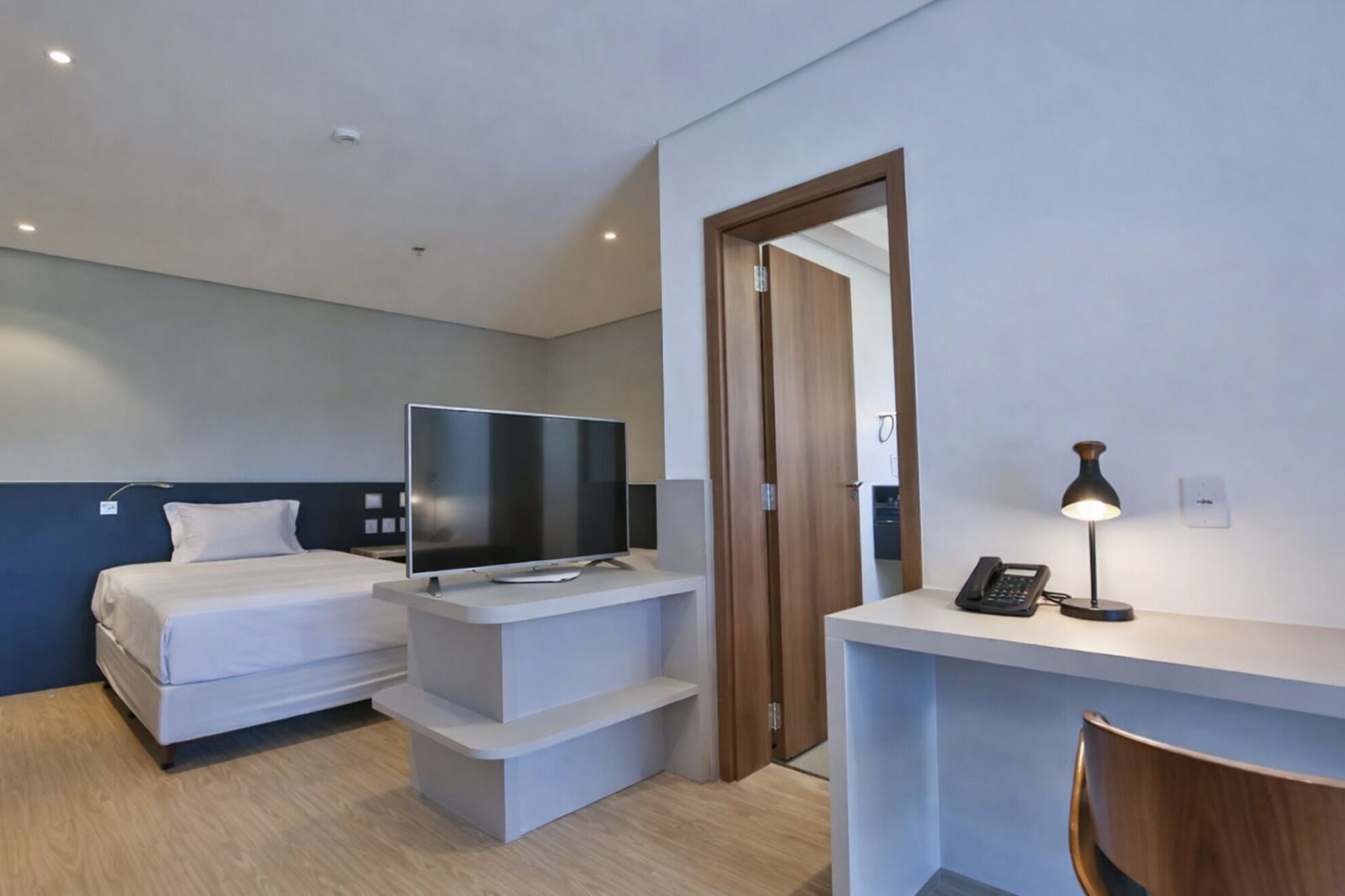 A modern hotel room with a bed, a TV on a stand, a desk with a phone, and a chair.