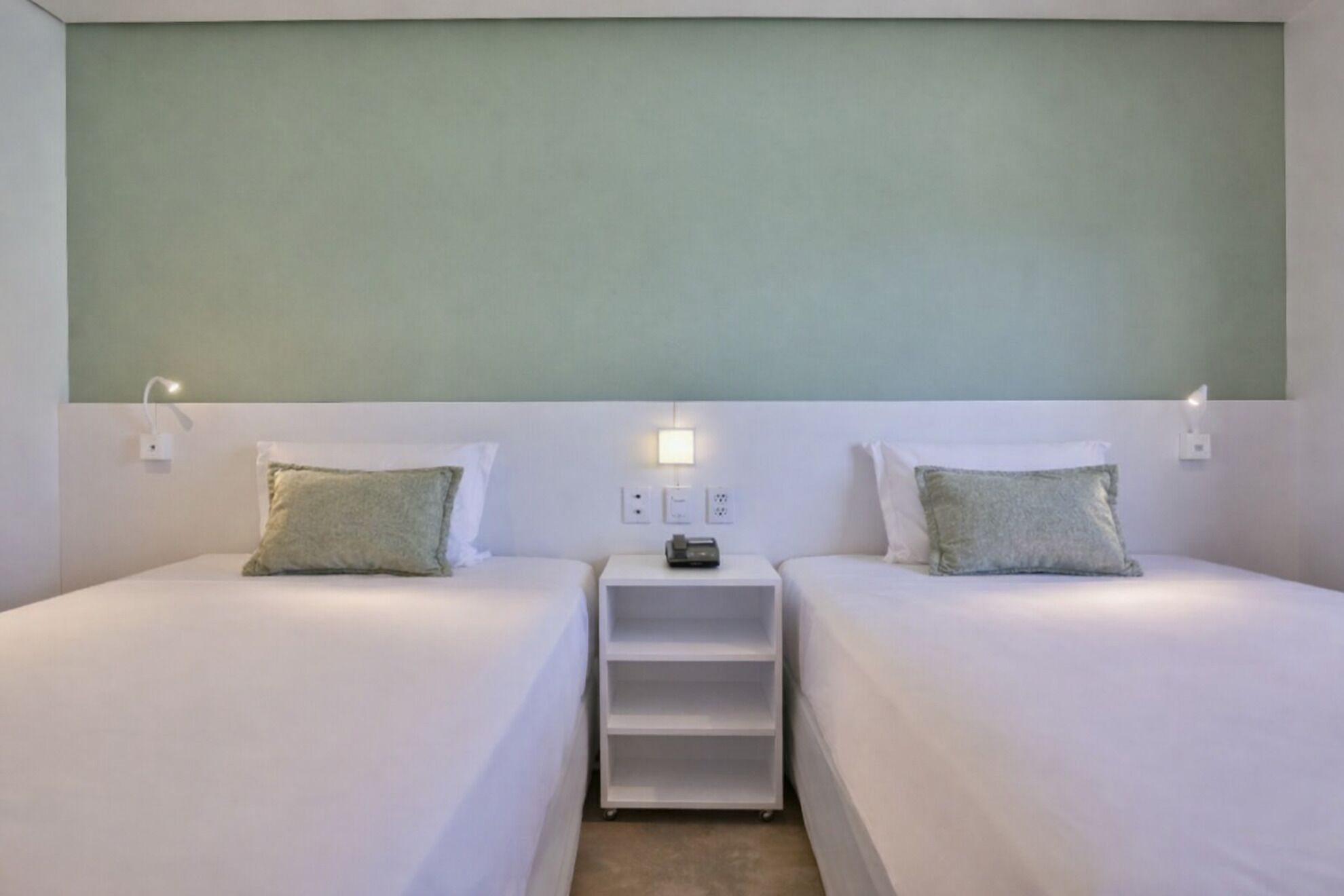 A hotel room with two single beds, a nightstand between them, and a wall-mounted shelf.