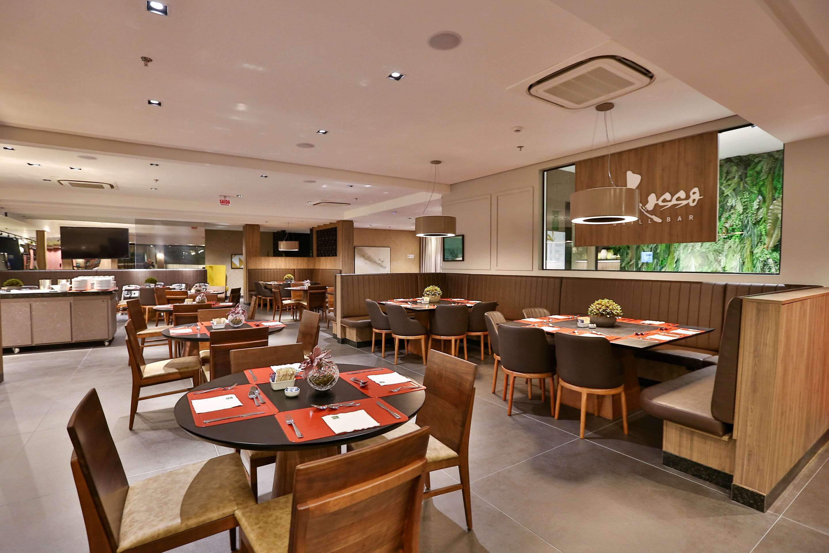 A modern restaurant with round tables, booths, and a large TV screen.