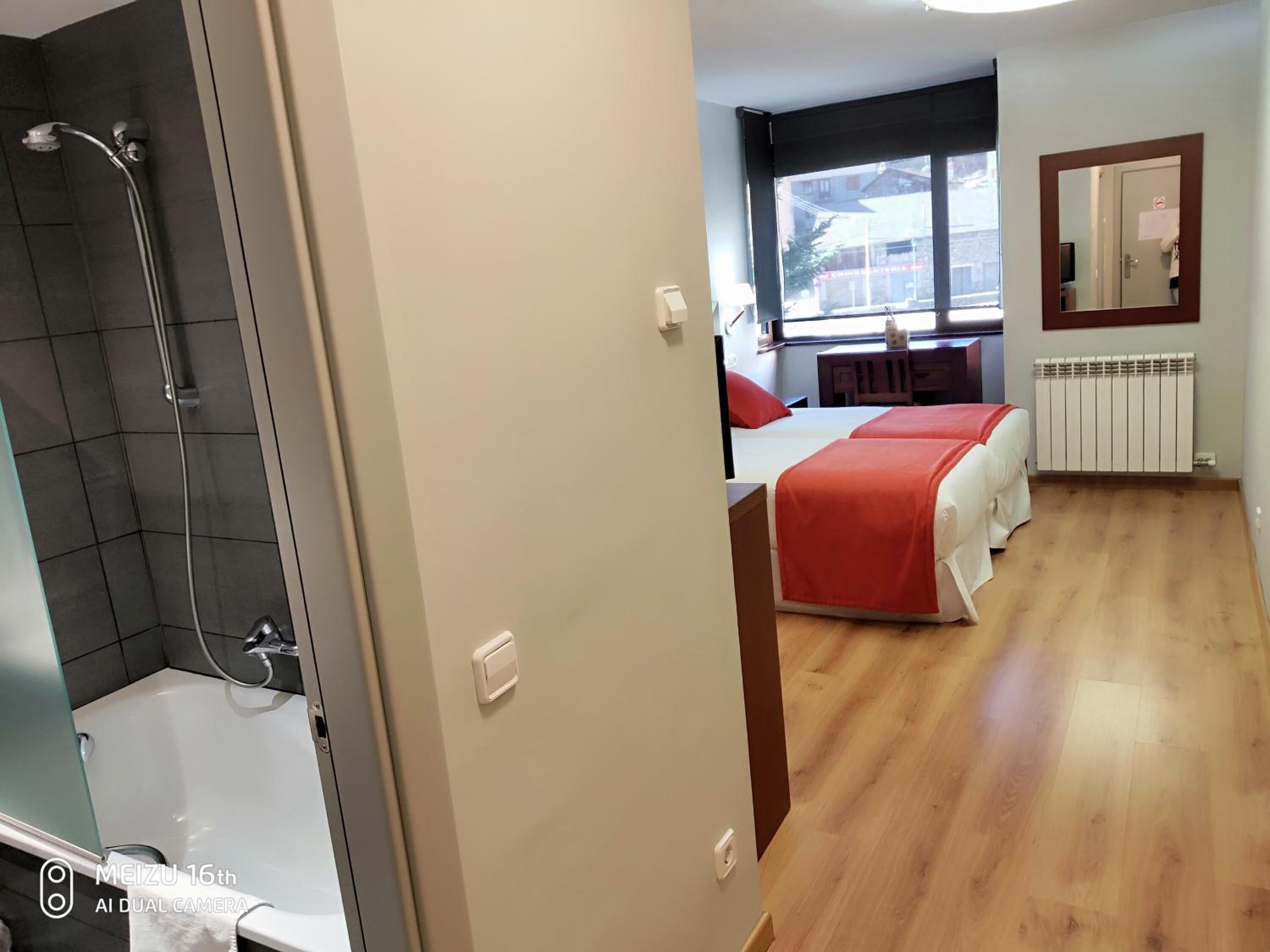A hotel room with a shower, a bed with a red blanket, a window with a view, and a radiator.