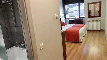 A hotel room with a shower, a bed with a red blanket, a window with a view, and a radiator.