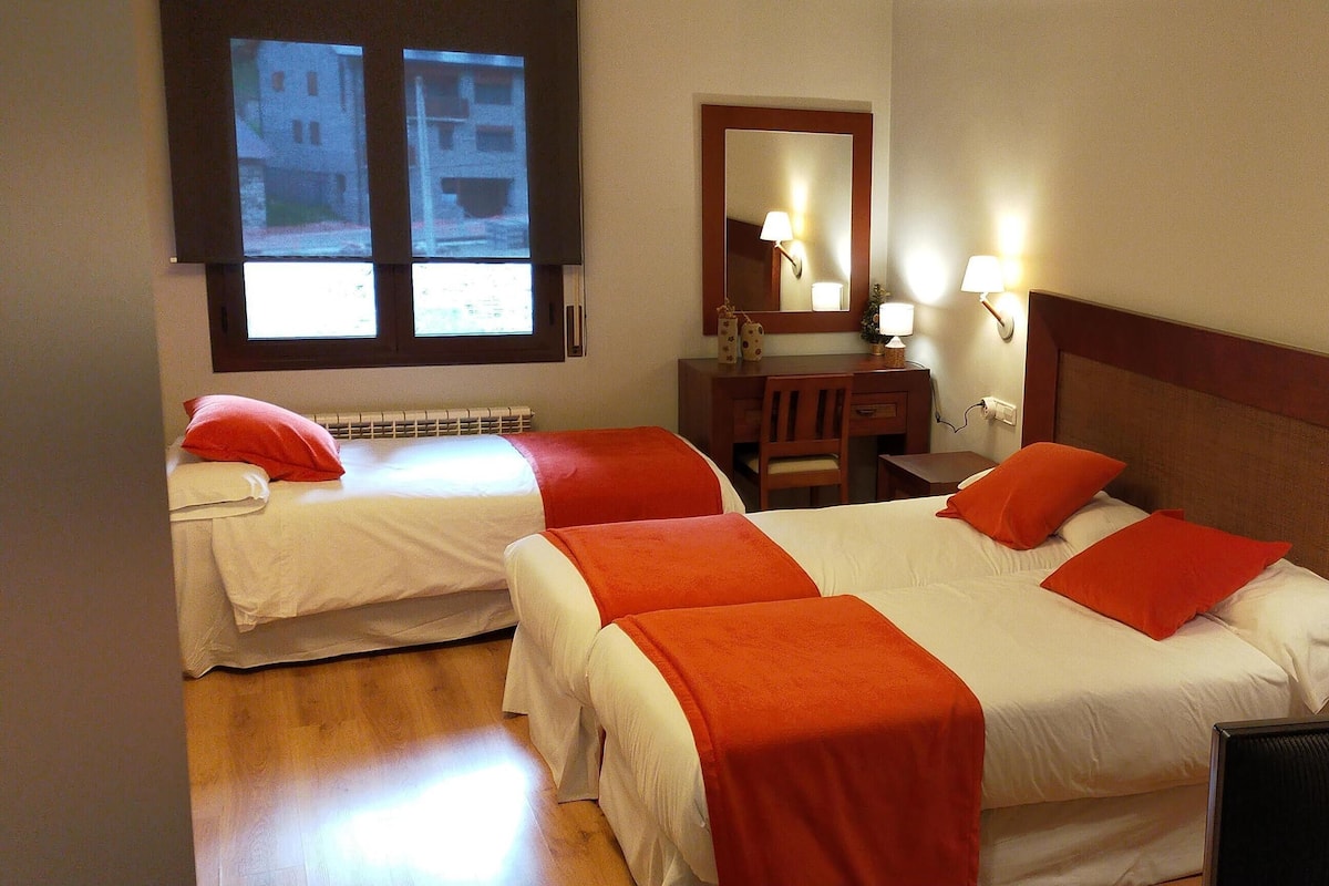 A hotel room with two beds, each with red pillows and white bedding. There is a wooden dresser with a mirror and two wall-mounted lamps.