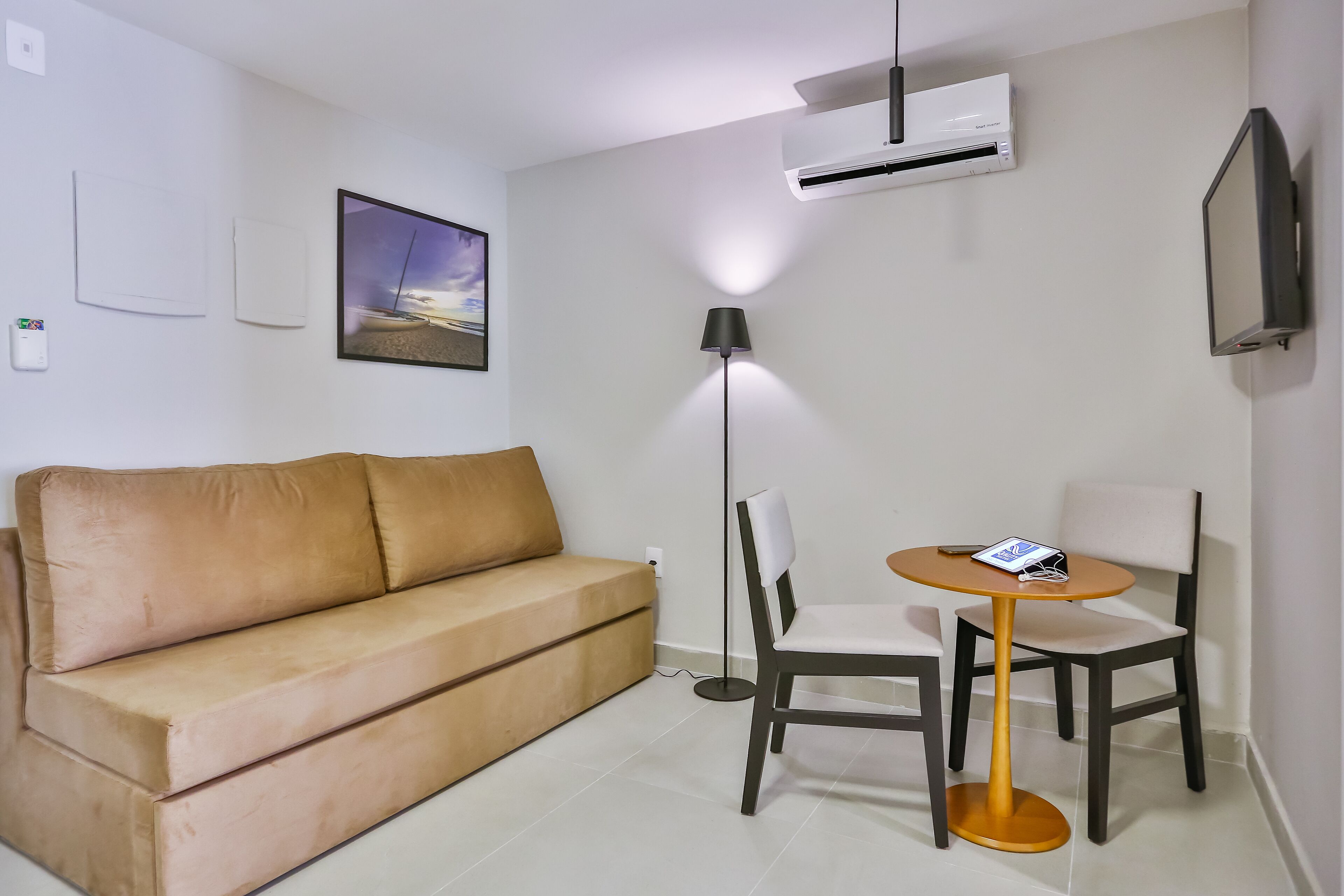 A room with a beige sofa, a small round table with chairs, an air conditioner, a floor lamp, and a framed picture on the wall.