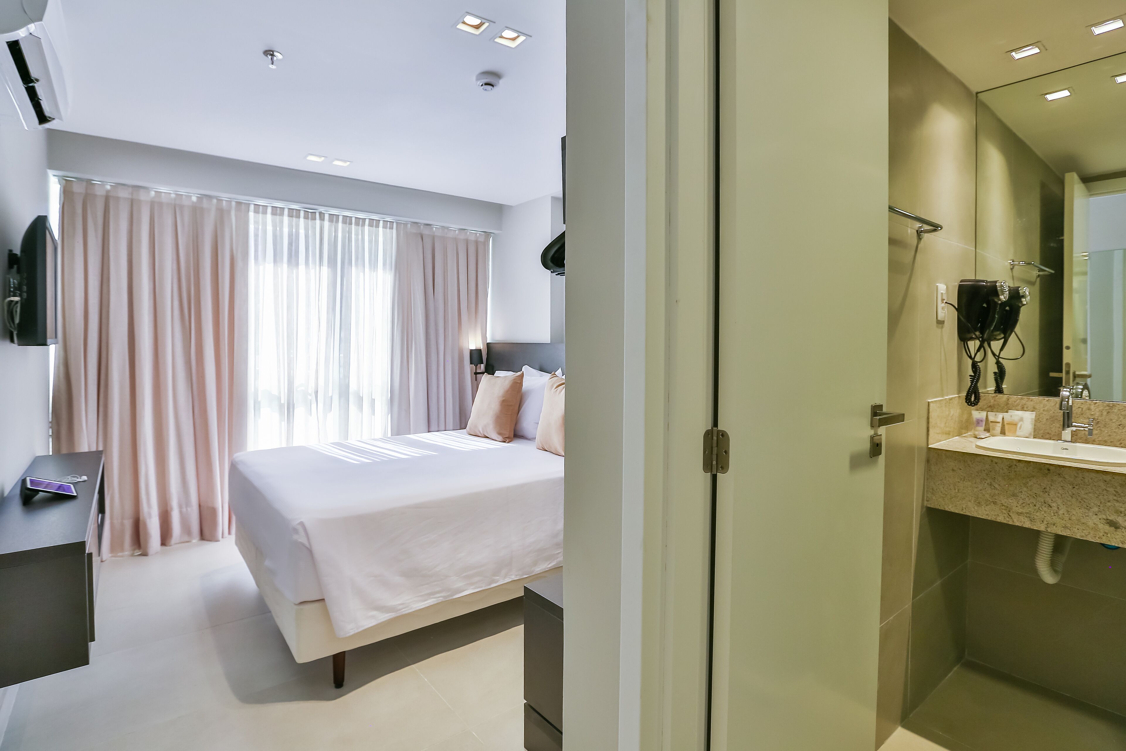 Una habitación de hotel moderna con cama, mesita de noche, televisión y un gran ventanal con cortinas.