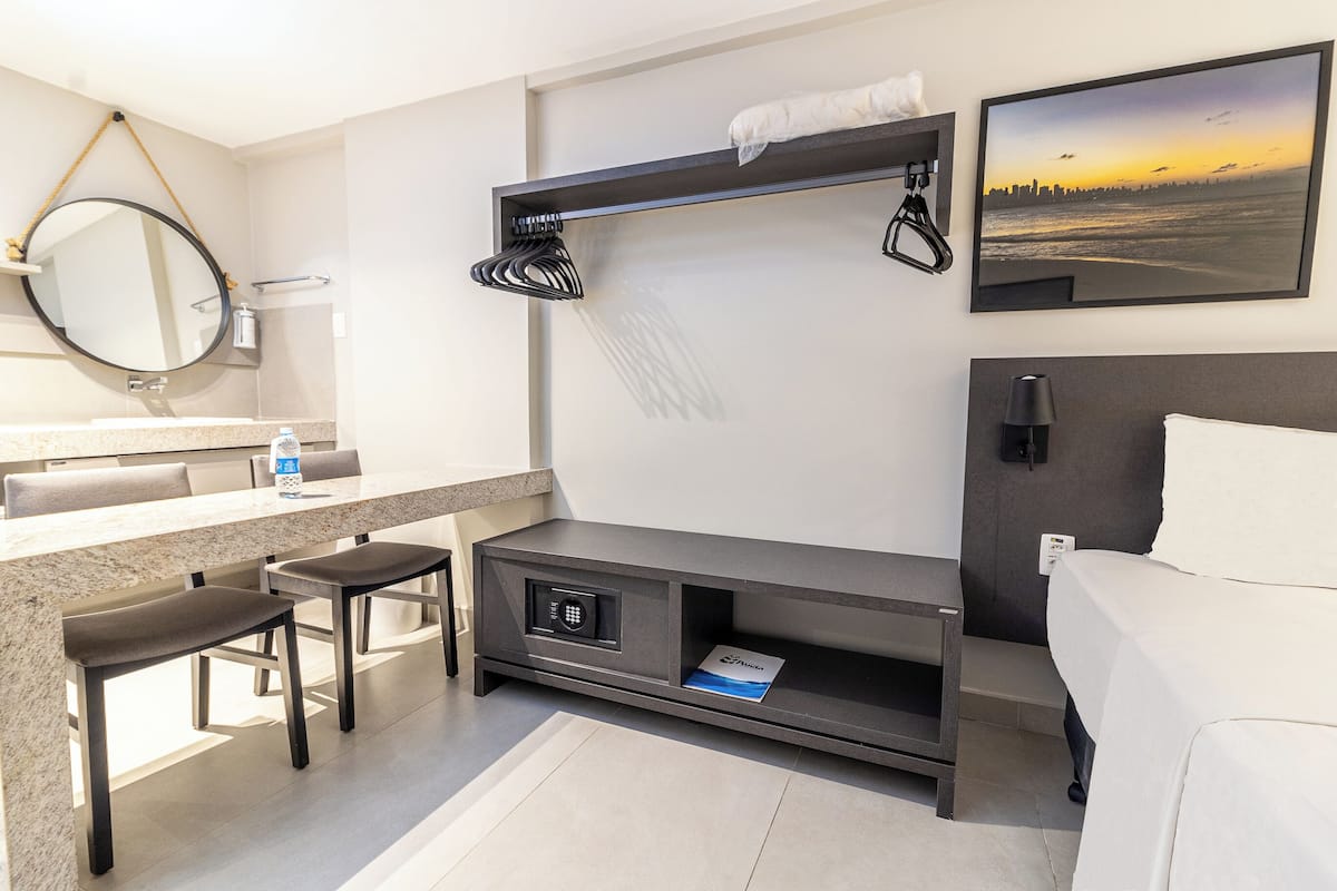 A modern hotel room with a large bed, a built-in desk with a chair, a vanity with a mirror, and a wall-mounted TV.