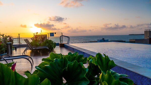 An infinity pool with a view of the sunset over the ocean, lounge chairs, and potted plants. - Quality Suítes João Pessoa (João Pessoa)