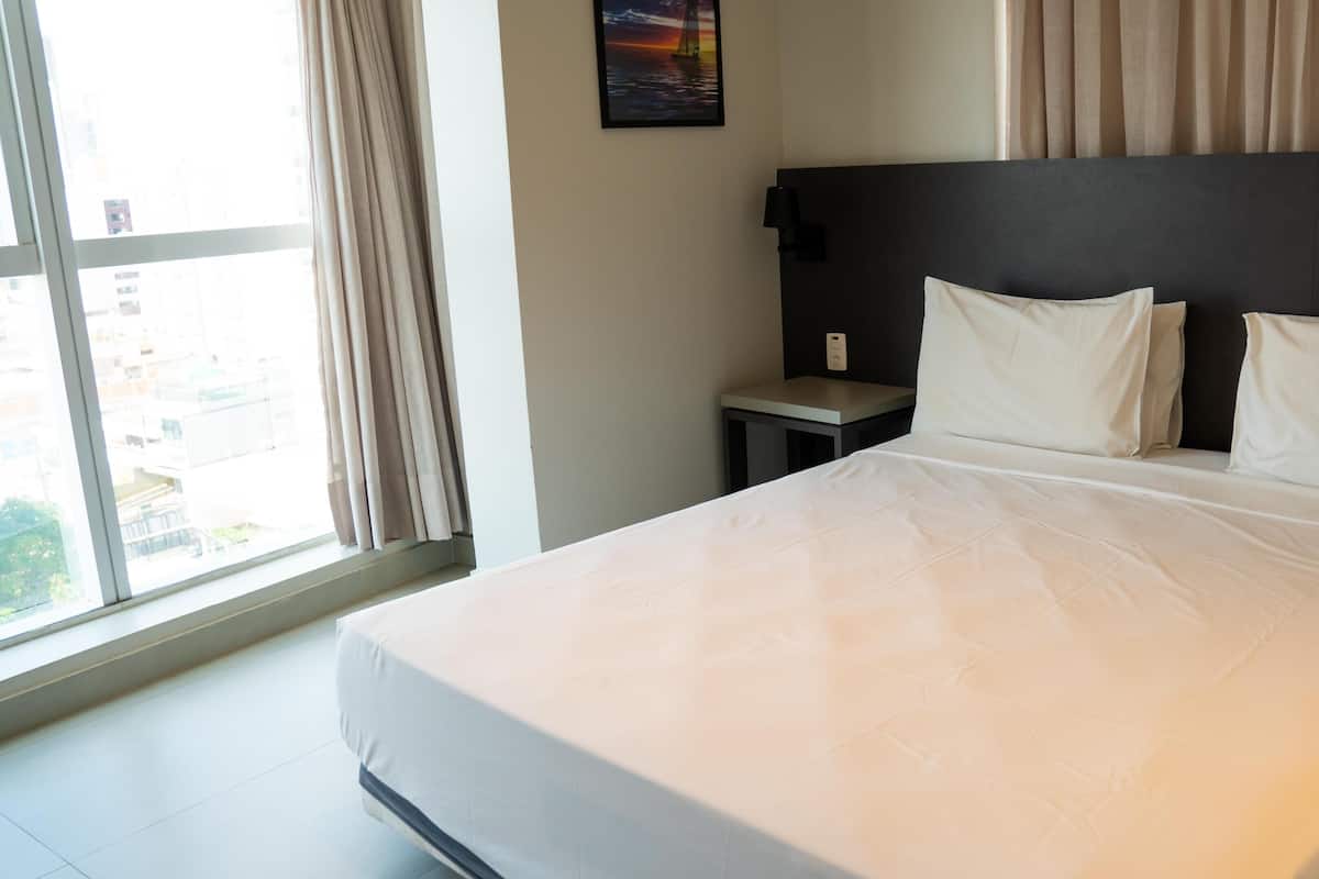 A hotel room with a large bed, a nightstand, a wall-mounted lamp, and a window with curtains.