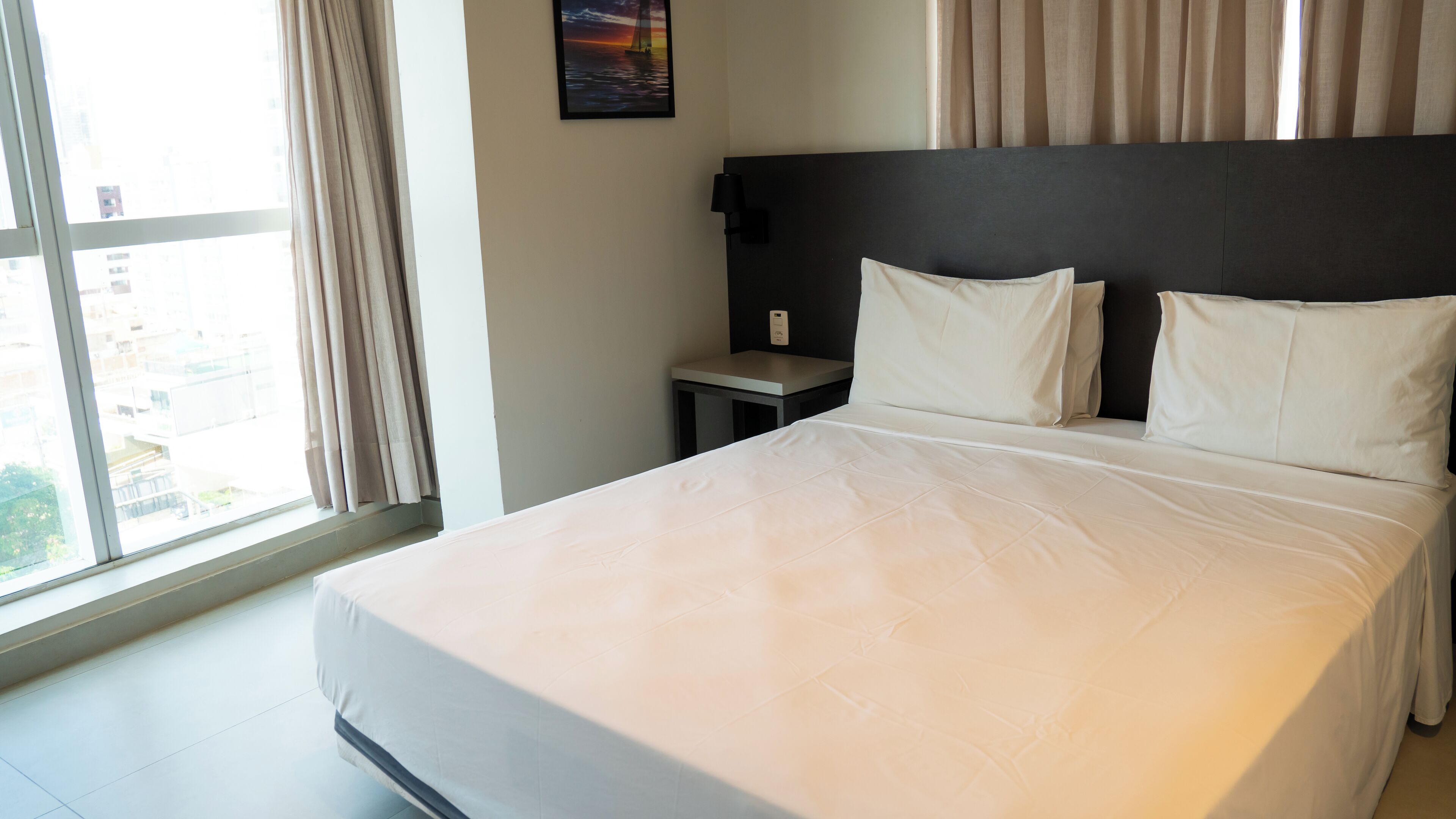 A hotel room with a large bed, a nightstand, a wall-mounted lamp, and a window with curtains.