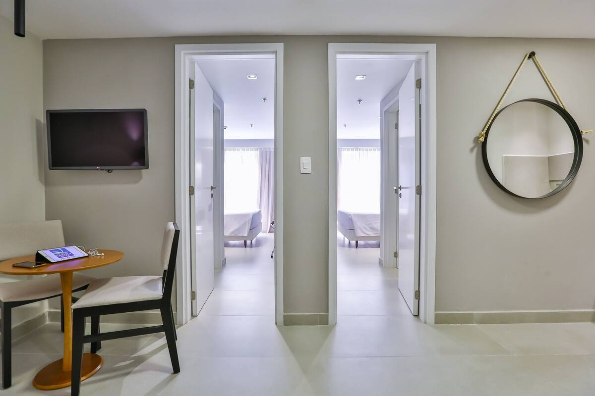 A hotel room with a flat-screen TV, a round mirror, a small table with a chair, and a bed with a white bedspread.