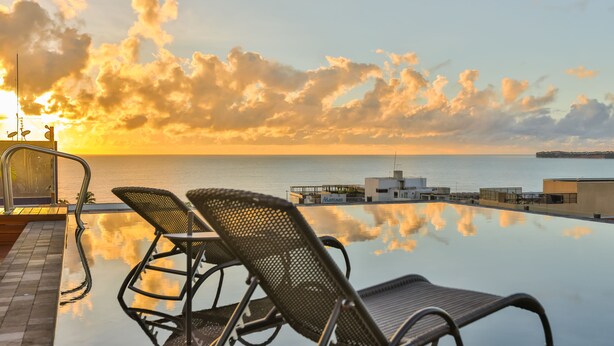 Piscina infinita com duas espreguiçadeiras e vista para o mar ao pôr do sol.