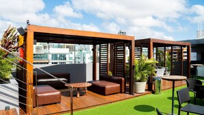 Terraço no telhado com mobiliário de madeira, grama sintética verde e vista para a paisagem urbana.