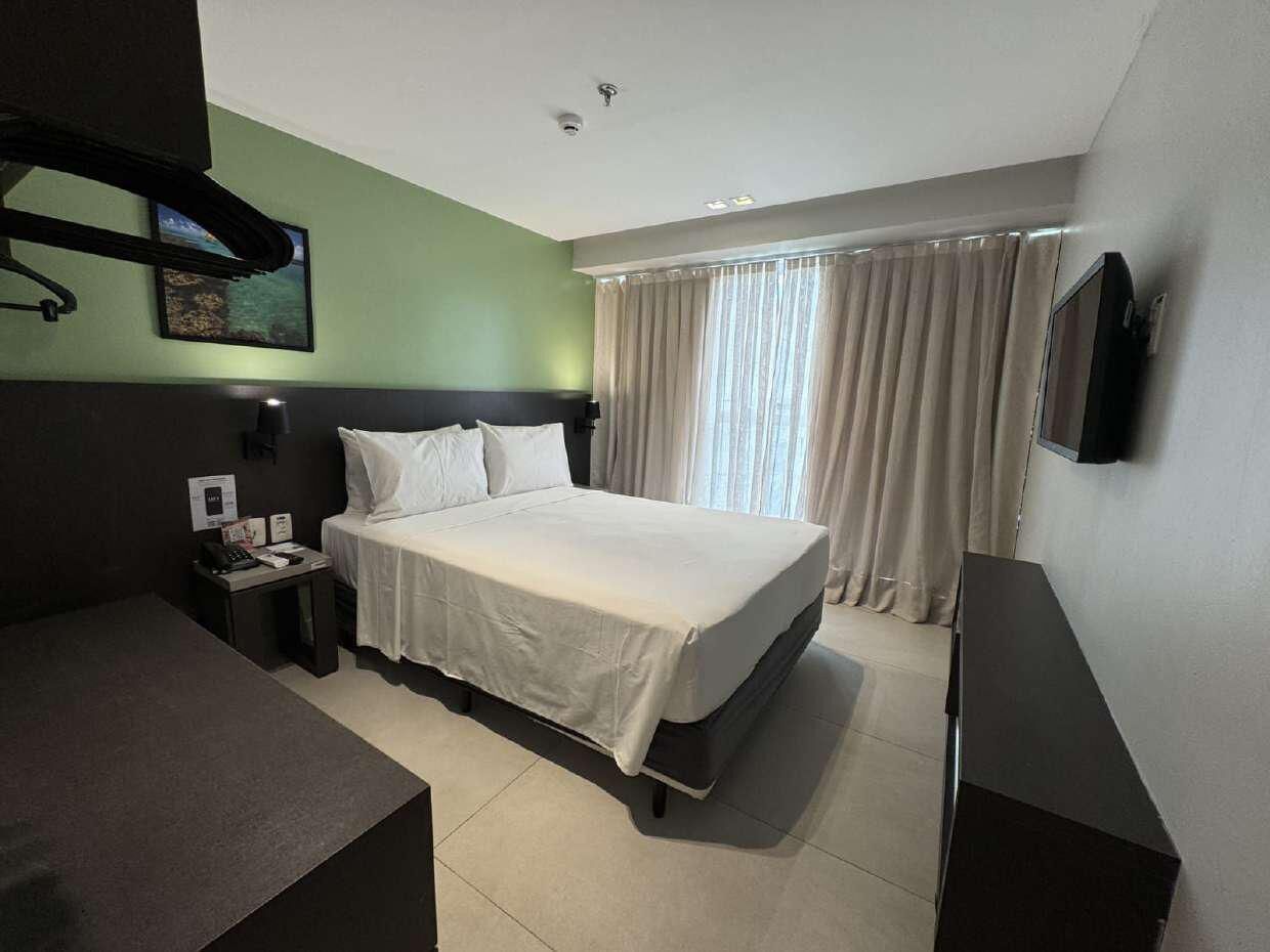 A hotel room with a bed, a desk, a chair, a TV, and a window with curtains.